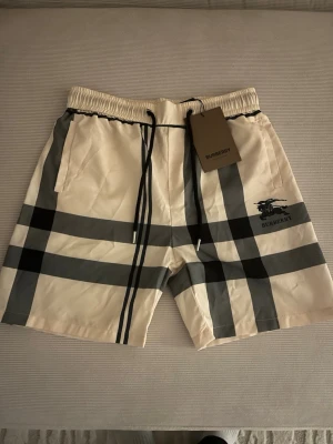 Beige rutiga shorts från Burberry - Snygga beige shorts från Burberry med klassiskt rutigt mönster i svart och grått. Shortsen har elastisk midja med snörning och sidofickor. Perfekta för en trendig och avslappnad stil. Dem är små i storlek så sitter bättre som M. Hör av er vid minsta lilla fråga. 
