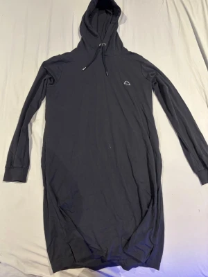 Mörkgrå hoodieklänning med huva - Långärmad mörkgrå hoodieklänning med huva och dragsko. Liten vit broderad detalj på bröstet. Mjuk bomullskänsla och ribbade muddar vid ärmslut och nederkant. Perfekt för en avslappnad och trendig look.