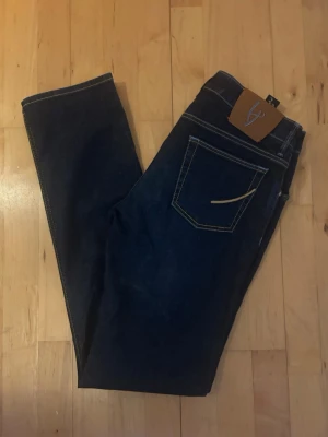 Mörkblå jeans från Handpicked Ravello - Snygga mörkblå jeans från Handpicked med kontrastsömmar och klassisk femficksmodell. Jeansen har knappgylf och detaljer i silverfärg. Materialet är kraftig denim som ger en tidlös look.