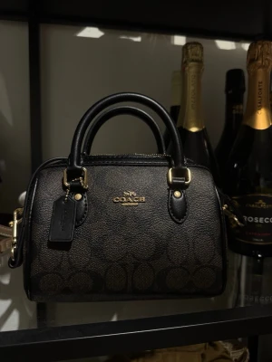 Coach Mini rowan handväska - Snygg handväska från Coach i brunt och svart med klassiskt monogrammönster. Väskan har två handtag, avtagbar axelrem och guldfärgade metalldetaljer. Tillverkad i tåligt lädermaterial och perfekt för dig som gillar stilrena accessoarer. 