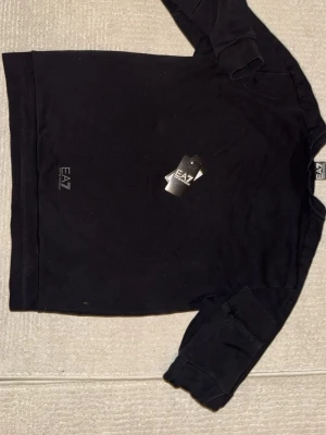 Svart långärmad topp från EA7 - Svart långärmad topp från EA7 Emporio Armani med stor logga framtill. Klassisk rund halsringning och mjukt material som känns skönt mot huden. Perfekt för en stilren och sportig look.