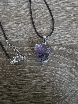 Halsband med hjärthänge och lila blomma - Unikt halsband med svart läderrem och ett hjärtformat hänge i transparent resin. Hänget innehåller en lila torkad blomma och en liten  fjäril, vilket ger en naturinspirerad och romantisk känsla. Perfekt accessoar för dig som gillar boho-stil.