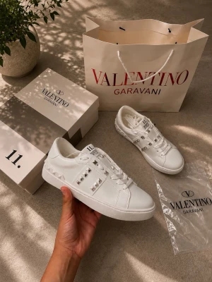 Vita Valentino Garavani sneakers - Snygga vita sneakers från Valentino Garavani med ikoniska nitar längs sidorna och på hälen. Skorna är låga och har en stilren design i vitt läder med matchande vita skosnören och detaljer. Perfekta för dig som vill ha en exklusiv touch på din outfit.
