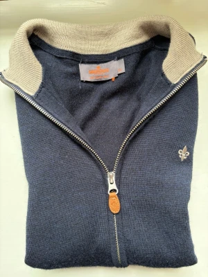Marinblå Morris fullzip - Mörkblå stickad fullziptröja med beige insida på kragen och en liten broderad detalj på bröstet. Tröjan har hel dragkedja framtill, ribbade muddar och en klassisk, stilren look. Perfekt för lager-på-lager och passar till det mesta.