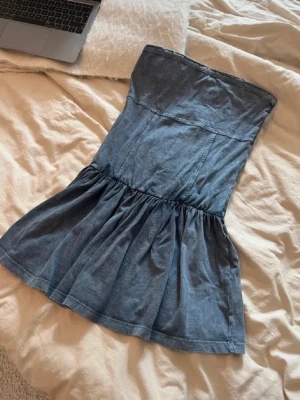 Axelbandslös jeansklänning - Säljer en snygg axelbandslös kort jeansklänning i blått. Klänningen har en figurnära överdel och utställd kjol med rynkade detaljer. Perfekt för dig som gillar en avslappnad men trendig stil.