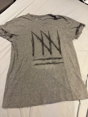 Grå t-shirt med grafiskt tryck - Säljer en grå t-shirt med korta ärmar och ett stort grafiskt tryck framtill i svart. T-shirten har en avslappnad passform och är tillverkad i mjukt bomullsmaterial. Perfekt för en chill och stilren look.