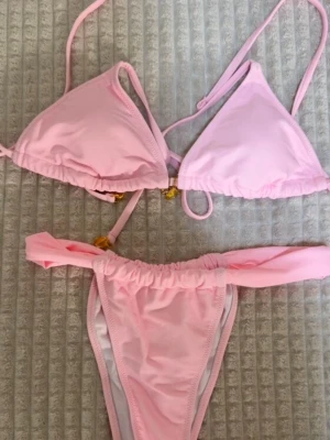 Ljusrosa bikini med knyt - Supersöt ljusrosa bikini med trekants-bh och knyt i sidorna. Bikinin har små gulddetaljer på banden och är perfekt för sommaren. Klassisk och enkel design som passar till strandhäng och pooldagar.