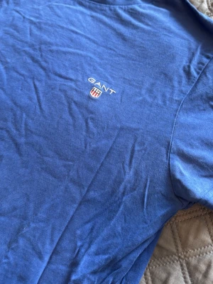 Blå t-shirt från GANT - Säljer en klassisk blå t-shirt från GANT med liten broderad logga och sköld på bröstet. T-shirten har rund halsringning och korta ärmar. Perfekt basic-plagg till garderoben.