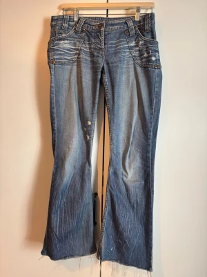 Blå bootcut jeans med slitningar - Säljer ett par blå bootcut jeans med slitna detaljer och råa kanter nertill. Jeansen har fem fickor, låg midja och snygga slitningar både fram och bak. Tillverkade i klassisk denim med en avslappnad vibe.