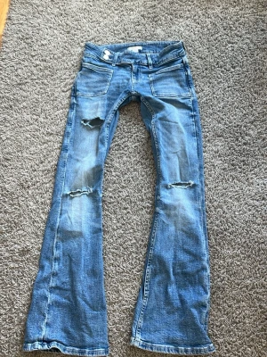 Blå slitna bootcut jeans - Ett par ljusblå bootcut jeans med slitningar och hål på båda benen. Jeansen har klassiska fem fickor och råa kanter längst ner. Perfekta för en avslappnad och trendig look.