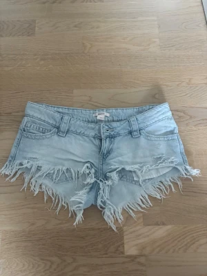 Ljusblå jeansshorts med fransar - Säljer ett par ljusblå denimshorts med råa, fransiga kanter och klassisk femficksdesign. Shortsen har bälteshällor och knappgylf. Perfekta för varma dagar och ger en avslappnad vibe. Knappt använda.