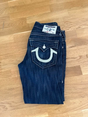 Mörkblå True Religion jeans - Säljer ett par mörkblå jeans från True Religion med kontrastsömmar och ikonisk fickdesign bak. Klassisk rak passform och detaljerade ficklock med knapp. Snygga att matcha med sneakers eller boots.