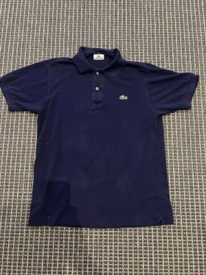 Marinblå pikétröja från Lacoste - Klassisk marinblå pikétröja från Lacoste med korta ärmar och krage. Tröjan har två knappar framtill och den ikoniska gröna krokodilloggan broderad på bröstet. Perfekt till jeans eller shorts för en clean och sportig look.