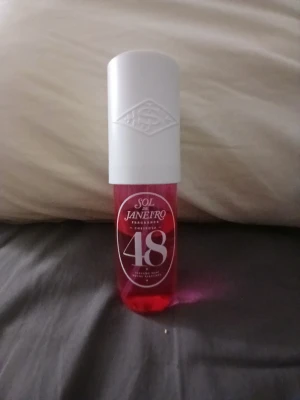 Sol de Janeiro Cheirosa 48 parfym mist - Sol de Janeiro Cheirosa 48 är en parfym mist i en cylinderformad flaska med vit kork och rosa transparent plast. Flaskan har en modern design med tydlig logga och siffran 48 på framsidan. Perfekt för dig som gillar fräscha och söta dofter.
