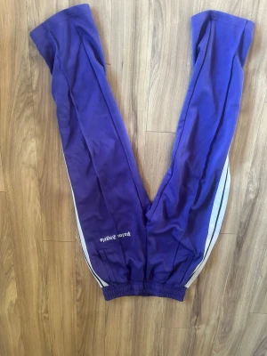 Lila Palm Angels dress - Säljer ett par lila mjukisbyxor från Palm Angels med vita sidoränder och logga tryckt på benet. Byxorna har resår i midjan och vid bensluten, samt raka ben och sportig look. Perfekta för en chill och trendig stil. samma med tröjan. hela dressen kostar 5000 ny. unik färg som inte säljs längre heller. storlek S 170-180cm