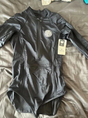   Rip Curl baddräkt  - Snygg svart långärmad baddräkt från Rip Curl med dragkedja framtill och rund hals. Tillverkad i stretchigt material med UPF50+ solskydd. Perfekt för surf eller strandhäng där du vill ha extra skydd mot solen. (helt oanvänd)