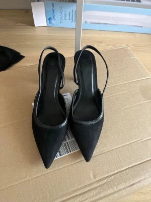 Svarta spetsiga klackskor - Säljer ett par svarta pumps med spetsig tå och smal klack. Skorna har öppen häl med slingback-rem och är tillverkade i mockaimitation med detaljer i slätt material. Perfekta för dig som gillar stilrena och eleganta skor.