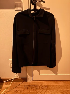 Svart overshirt med dragkedja - Stilren svart overshirt med rak passform och dragkedja framtill. Två stora bröstfickor med lock ger en cool utility-vibe. Tillverkad i ett mjukt tyg som känns bekvämt och passar perfekt till lager-på-lager. Klassisk krage och långa ärmar.