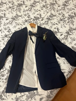Mörkblå kostymkavaj med skjorta, byxa och fluga. För barn - Snygg mörkblå kavaj med klassisk passform och matchande vit skjorta. Kavajen har två framfickor, knappstängning och en dekorativ blomma på slaget. Perfekt för student eller bal. Skjortan är långärmad och har knappar hela vägen.