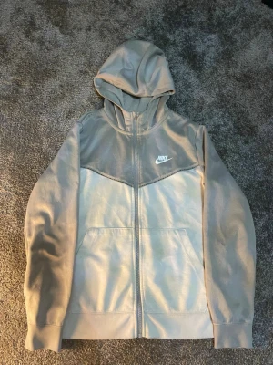 Grå zip hoodie från Nike - Snygg grå hoodie från Nike med dragkedja framtill och vit logga på bröstet. Tröjan har huva och två fickor framtill. Perfekt för en avslappnad och sportig stil. Tillverkad i mjukt material som känns skönt mot huden.