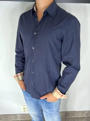 Burberry skjorta  - Stilren mörkblå skjorta med klassisk krage och knappar framtill. Manschetterna har snygga beige-rutiga detaljer som ger en unik touch. Skjortan är långärmad och har en normal passform, perfekt till jeans eller chinos.