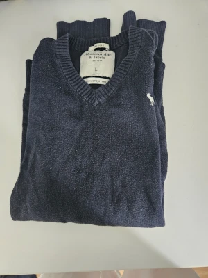 Mörkblå stickad tröja Abercrombie - Mörkblå stickad tröja från Abercrombie & Fitch i cashmereblandning. Tröjan har V-ringning, ribbade muddar och en liten vit logga broderad på bröstet. Perfekt för en stilren och avslappnad look. Passar (M)