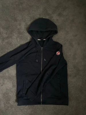 Mörkblå hoodie med dragkedja Colmar - Snygg mörkblå hoodie från Colmar med dragkedja framtill och två fickor. Klassisk huva med dragsko och ett broderat Colmar-märke på bröstet. Perfekt för en avslappnad och sportig stil. Tillverkad i mjukt material för extra komfort.