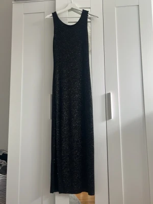 Svart långklänning med glitter - Snygg svart långklänning med glittriga prickar och ärmlös design. Klänningen har en rak och enkel siluett som ger ett stilrent intryck med slits nedtill. Perfekt för dig som vill sticka ut med lite extra glitter. Materialet känns mjukt och följsamt. Står igen storlek men skulle säga att klänningen motsvarar en S/M