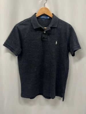 Mörkblå piké från Polo Ralph Lauren - Klassisk mörkblå pikétröja från Polo Ralph Lauren med vit broderad logga på bröstet. Tröjan har korta ärmar, krage och knappar vid halsen. Slim fit-modell i mjuk bomull som passar perfekt till jeans eller shorts.