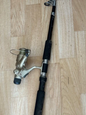 Svart fiskespö med rulle - Säljer ett svart fiskespö med tillhörande rulle. Spöet har svarta grepp i skumgummi och detaljer i silver. Rullen är i metallfärg och sitter monterad på spöet. Perfekt för fiskeäventyr vid sjö eller hav.