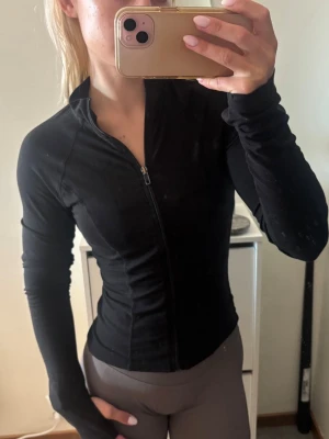 Svart sporttröja med dragkedja - Säljer en svart sporttröja med hel dragkedja framtill och hög krage. Jackan har lång ärm och är figurnära i modellen, perfekt för träning eller löpning. Materialet ser ut att vara stretchigt och följsamt för maximal komfort.