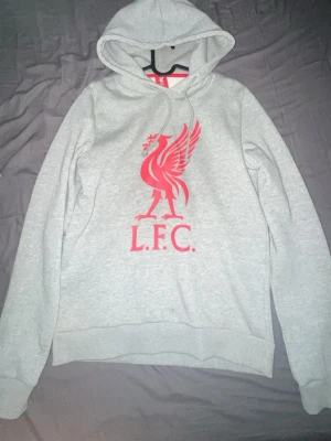 Grå Liverpool FC hoodie  - grå hoodie med  Liverpool FC-logga på bröstet. Tröjan har huva med dragsko och ribbade muddar vid ärmar och nederkant. Perfekt för dig som vill visa ditt stöd för LFC på ett stilrent sätt.