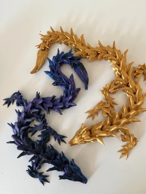 Drakformade fidget accessoarer i blått och guld - Unika 3D-printade fidget-leksaker och kinetiska skrivbordsdekorationer, tillverkade i högkvalitativ PLA-plast. Perfekta att ha på skrivbordet hemma eller på kontoret för att minska stress, hålla händerna sysselsatta eller bara som en snygg inredningsdetalj.  ✔ Passar både barn och vuxna ✔ Perfekta som present eller samlarobjekt ✔ Finns i flera modeller och färger