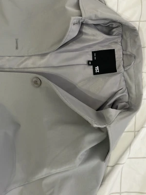 Ljusgrå trenchcoat från 157 - Stilren ljusgrå trenchcoat från 157 med klassisk krage, knäppning framtill och matchande bälte i midjan. Jackan har släta fickor och är helfodrad för extra komfort. Perfekt för dig som gillar minimalistisk och modern stil.
