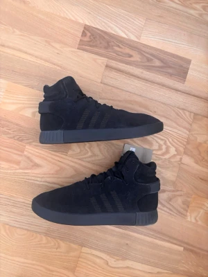 Adidas Tubular Invader Svart Sneakers Strl 42 - Snygga helsvarta sneakers med högt skaft och klassisk siluett. Skorna har en ovandel i mocka och diskreta ränder på sidan. Tjock sula och vadderad häl ger extra komfort. Perfekta för dig som gillar stilrena och minimalistiska skor.