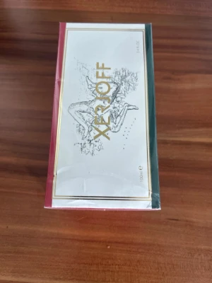 Xerjoff parfym 100ml - Lyxig parfym från Xerjoff i en elegant rektangulär förpackning med gulddetaljer och konstnärlig illustration på framsidan. Kartongen har färgade kanter i rött och grönt samt vit bas. Flaskan rymmer 100 ml.