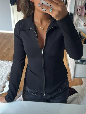 Svart långärmad topp med dragkedja - Snygg svart långärmad topp från zara med hel dragkedja framtill och krage. Figurnära passform som ger en clean och stilren look. Perfekt att matcha med jeans eller kjol för en trendig outfit. Använd endast en gång.
