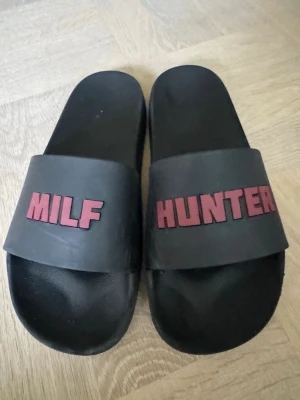 Svarta slides/tofflor - Milf Hunter  - Svarta sandaler i slides-modell med breda remmar där det står 'MILF' på ena och 'HUNTER' på andra i rödrosa bokstäver. Tillverkade i syntetmaterial och har en klassisk, enkel form som passar till avslappnade outfits.