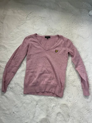 Ljusrosa stickad tröja Lyle & Scott - Ljusrosa stickad tröja från Lyle & Scott med v-ringning och gul logga på bröstet. Tröjan har långa ärmar och ribbade muddar vid ärmslut och nederkant. Perfekt för dig som gillar en enkel men snygg stil. Andvänd 2 ggr