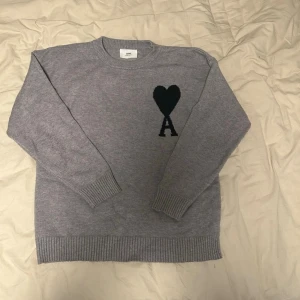 Ami paris sweatshirt  - Säljer en stilren grå stickad tröja med svart hjärta och bokstaven A på bröstet. Tröjan har rund halsringning, långa ärmar och ribbade muddar vid ärmslut och nederkant. Perfekt för dig som gillar enkel men snygg design.