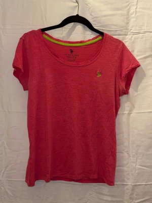 Rosa t-shirt från U.S. Polo Assn. - Snygg rosa t-shirt från U.S. Polo Assn. med klassisk rund halsringning och liten broderad logga i gult på bröstet. Perfekt för en avslappnad och stilren look. T-shirten har korta ärmar och är tillverkad i mjuk bomull. Den är lite mindre i  storleken skulle säga att den passar m/l