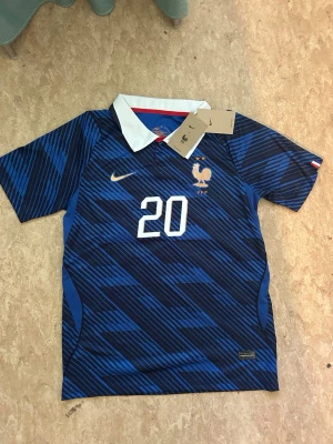 Blå Frankrike fotbollströja Nike - Säljer en blå fotbollströja från Nike med Frankrikes landslagsemblem och nummer 20. Tröjan har ett mönster i olika blå nyanser, vit krage och korta ärmar. Perfekt för dig som älskar fotboll och vill stötta Frankrike på matchen!