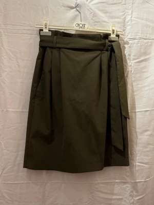 Olivgrön kjol med knytband - Stilren olivgrön kjol med knytband i midjan och rak passform. Kjolen har en enkel design och är perfekt för dig som gillar minimalistisk stil. Midjebandet ger en snygg detalj och gör att kjolen sitter bekvämt.