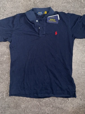 Marinblå pikétröja från Polo Ralph Lauren - Klassisk marinblå pikétröja från Polo Ralph Lauren med röd broderad logga på bröstet. Tröjan har krage, knappar vid halsen och korta ärmar. Perfekt för en stilren och avslappnad look.