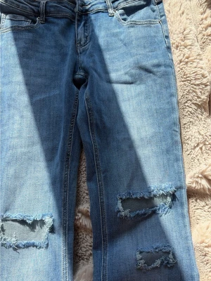 Ljusblå slitna bootcut jeans - Säljer ett par ljusblå jeans med bootcut passform och slitna detaljer på båda benen. Jeansen har klassisk femficksdesign och råa hål vid knäna för en trendig look. Perfekta för dig som gillar en avslappnad och cool stil.