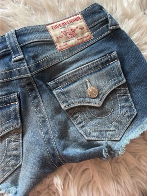 Blå jeansshorts från True Religion - Snygga blå jeansshorts från True Religion med råa kanter och klassiska bakfickor med lock och silverfärgade knappar. Shortsen har en avslappnad passform och coola slitningar för en trendig look.