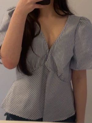 Randig v-ringad blus med puffärm - Säljer en snygg blus med smala vita och blå ränder. Blusen har djup v-ringning, puffiga korta ärmar och lite öppen på baksidan!