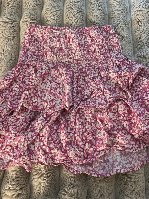 Rosa blommig volangkjol - Supersöt kort kjol med rosa och vitt blommönster och volangdetaljer. Kjolen har smock i midjan för en bekväm passform och är gjord i ett lätt, luftigt material som passar perfekt till varmare dagar.