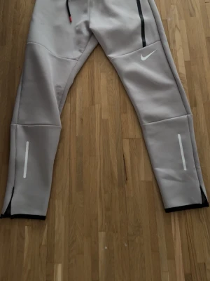Grå mjukisbyxor från Nike - Snygga grå mjukisbyxor från Nike med svarta och vita detaljer. Byxorna har snörning i midjan, vita reflexränder på benen och en svart linje längs ena fickan. Perfekta för chill eller träning.