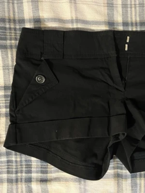 Svarta shorts från BAY med knappdetalj - Säljer ett par svarta shorts från BAY med snygg knappdetalj på fickan och diskret dragkedja framtill. Shortsen har en klassisk passform och är perfekta för varma dagar. Tillverkade i ett mjukt bomullsmaterial som känns skönt mot huden.